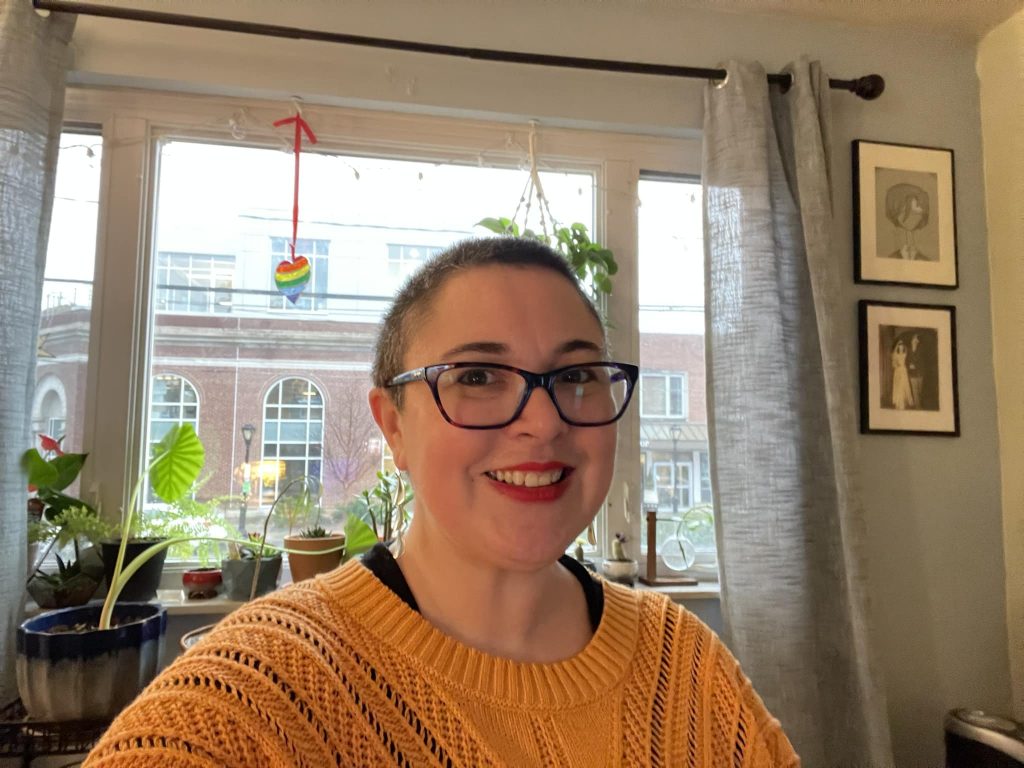 A white woman with a shaved head in glasses and a yellow sweater smiles in front of a window full of plants